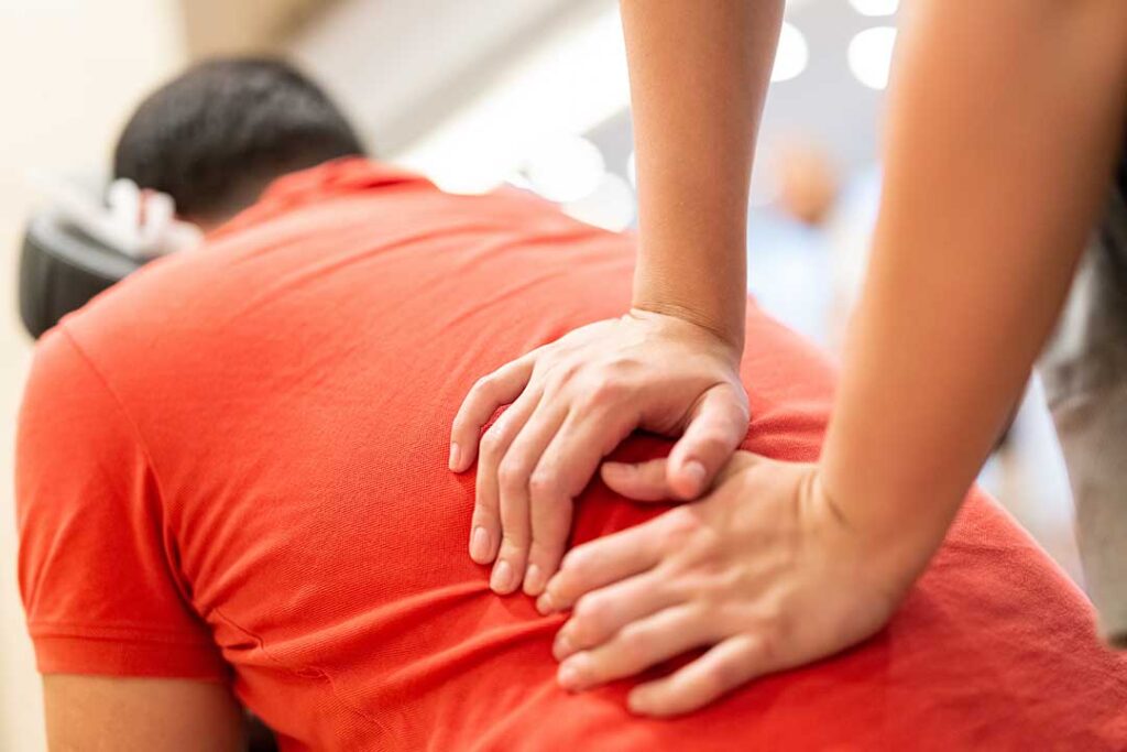 An adult sits indoors near a window on a rainy day, gently massaging their knee with a relaxed expression.